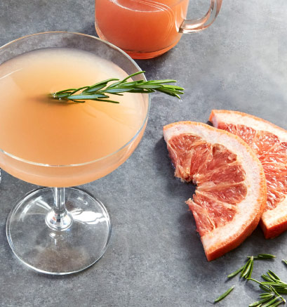 A cocktail glass filled with an orange drink and garnished with a sprig of rosemary surrounded by grapefruit slices, rosemary leaves, and a glass of juice